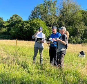 Three people standing at the side of a river, collecting habitat quality data on sheets.