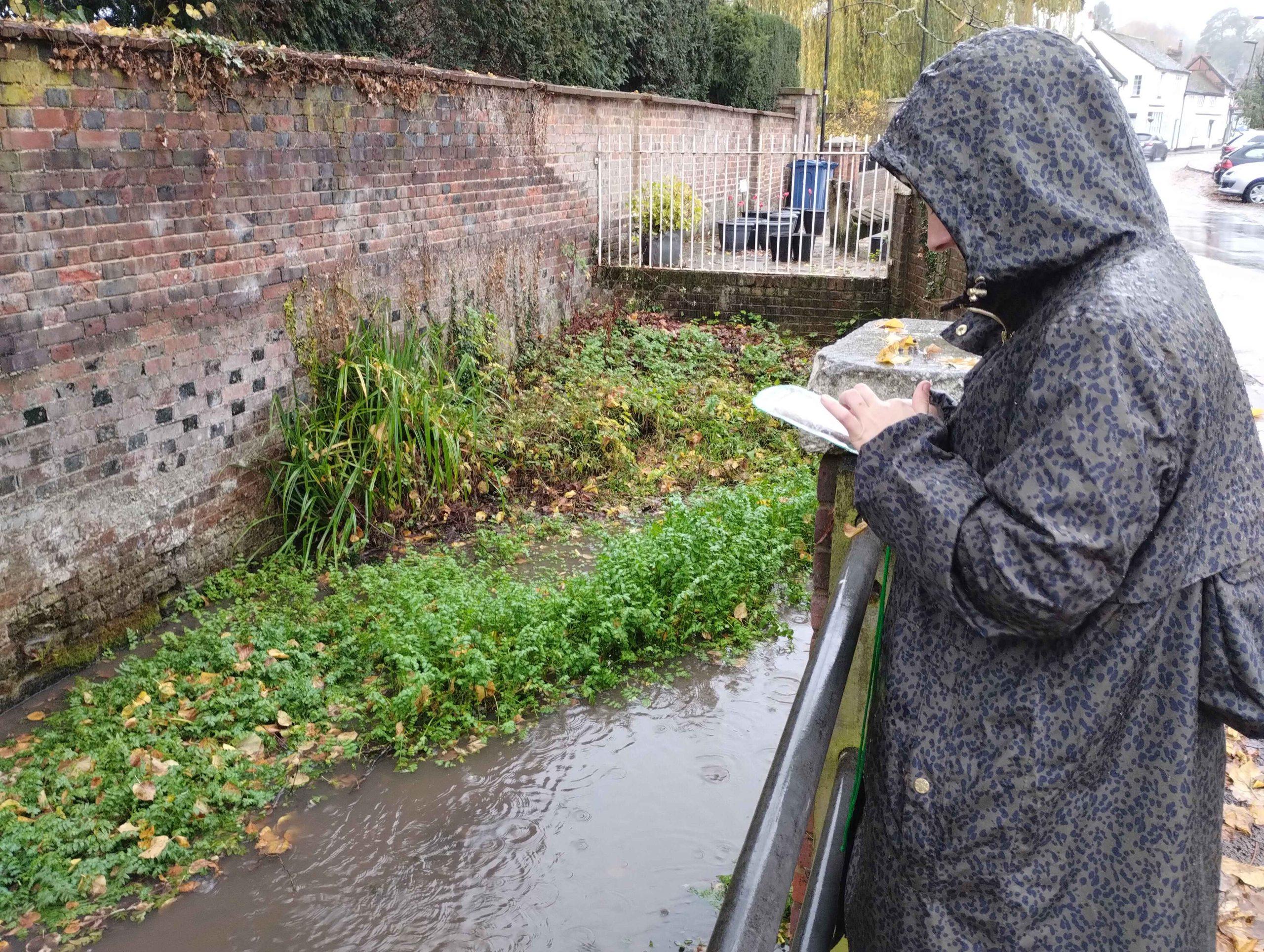 Photo of person collecting survey data by river in the rain.
